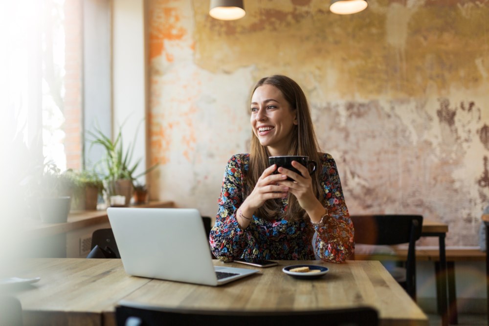 Jente holder en kopp kaffe og smiler foran PC-en.